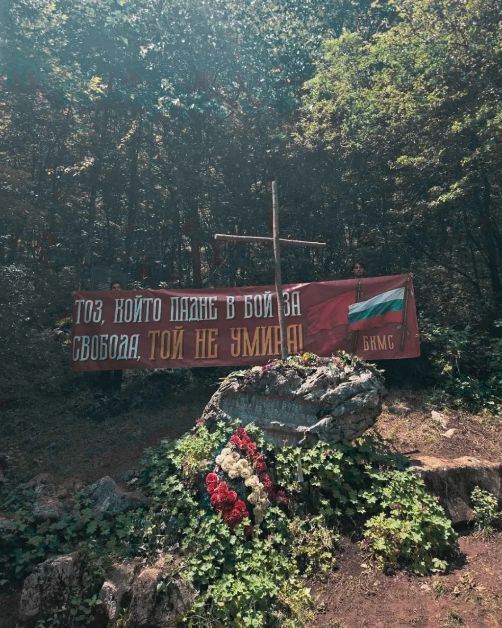 Транспарант на БКМС пред кръста и мемориалния камък, отрупан с цветя.