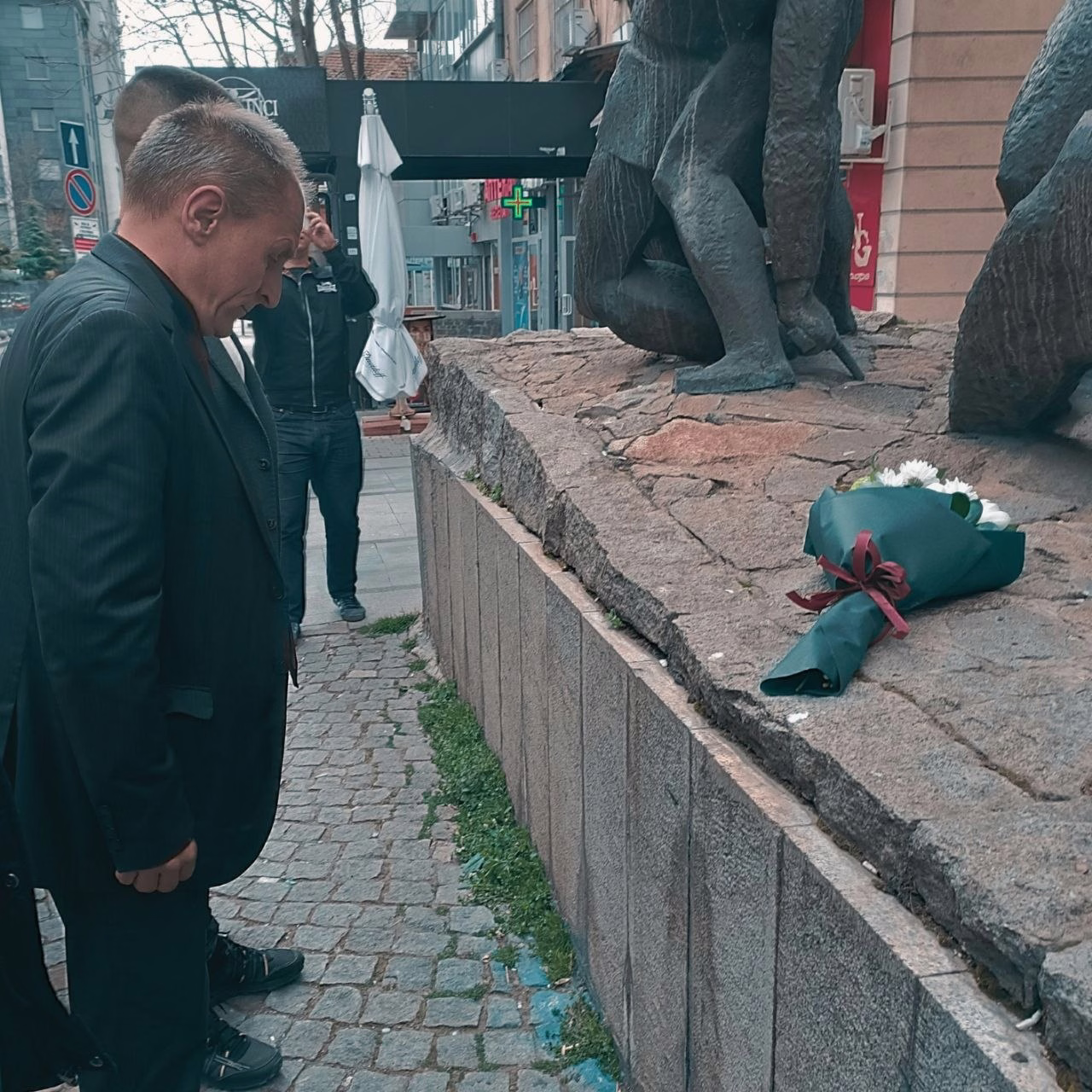 Членове на БКМС стоят пред паметник, върху който е положен букет цветя. Те са наведени напред в градска среда.