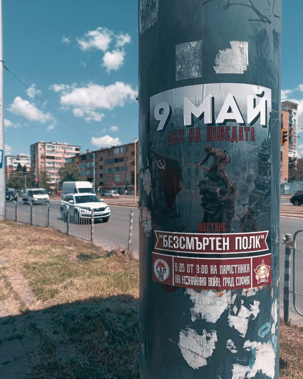 Плакат за шествието „Безсмъртен полк“, залепен на метален стълб край булевард в София. На изображението са представени членове на БКМС, които издигат знаме върху руини.