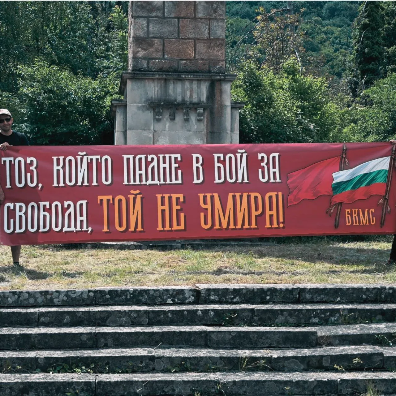 Членове на БКМС пред паметник в планинска местност. Група позира с червени знамена на фона на гора и каменист терен.