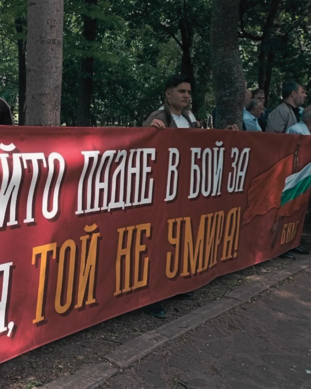 Членове на БКМС пред паметник в паркова среда. На заден план се виждат дървета и тревни площи.