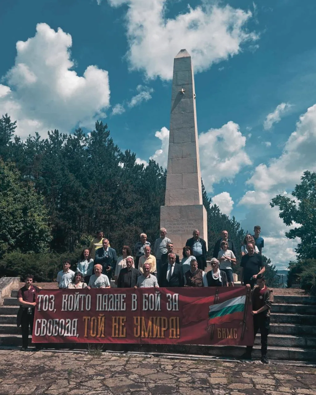 Групова снимка на участниците в честването пред каменен обелиск, с червено-бял транспарант на БКМС с надпис „Тоз, който падне в бой за свобода, той не умира!" и българското знаме.