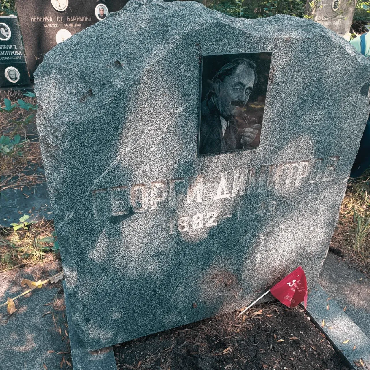Близък план на паметника на Георги Димитров с неговия портретен барелеф, надпис „Георги Димитров / 1882" и поставено малко червено знаме на БКМС, на фона на гробищни паметници и зеленина.